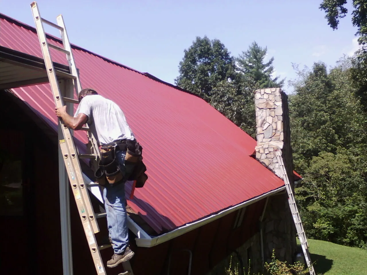 Roofer on a ladder beside a red metal roof and stone chimney — Soffit & Fascia Repair by Roofing Brothers in Logan